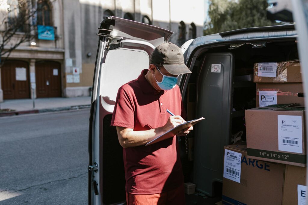 Courier with mask and clipboard arranging packages in delivery van.